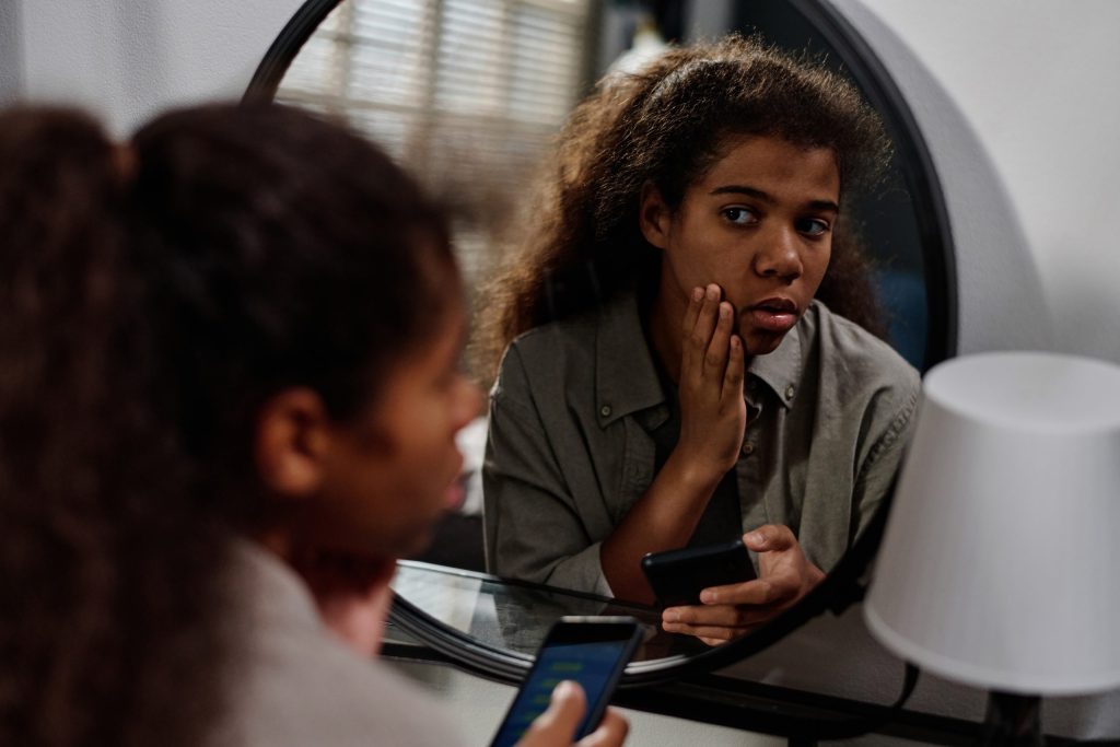 Effects of technology on mental health shown as young woman comparing herself in mirror while holding phone